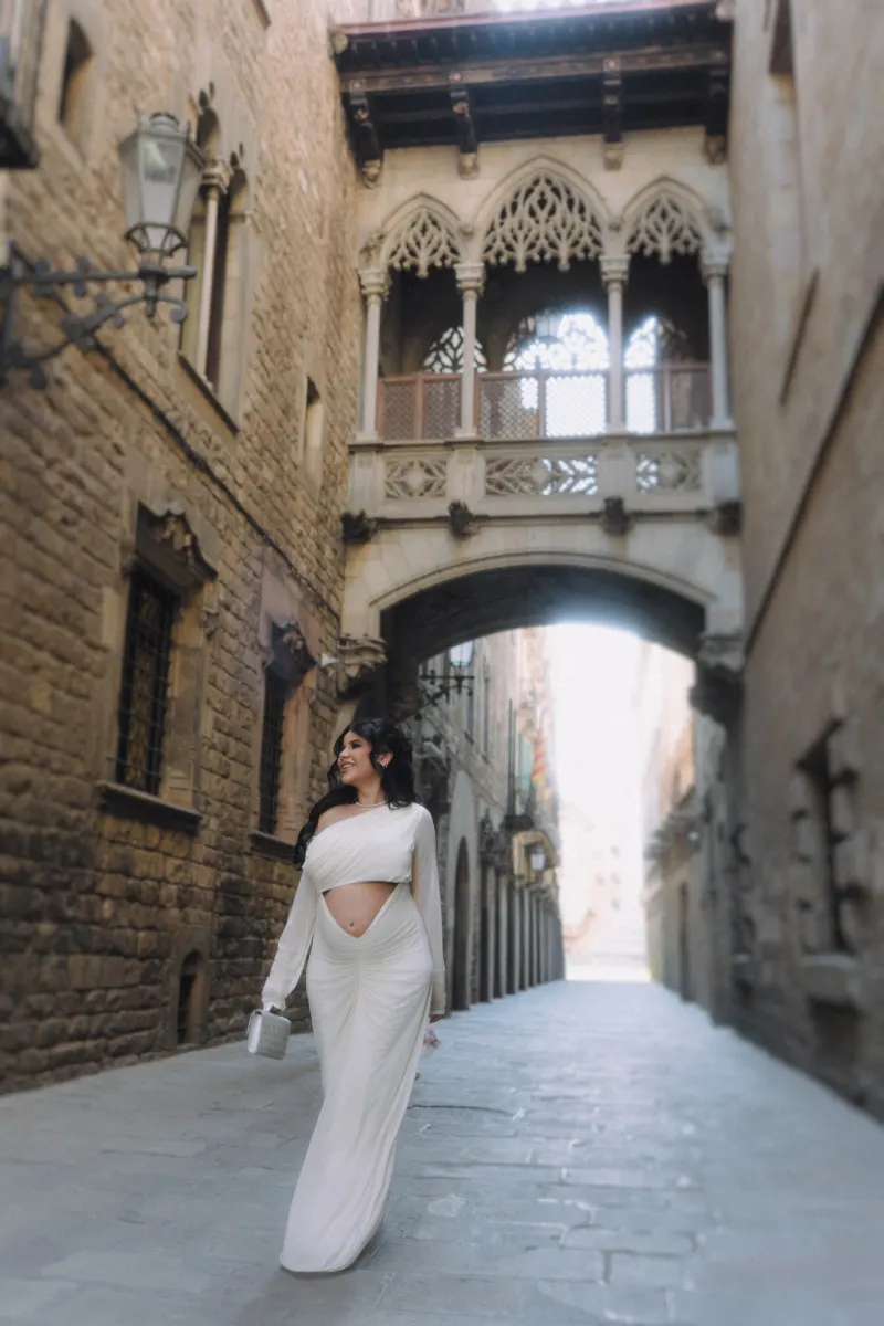 Smiling expectant mother in a white gown strolls through a historic city street during a Vacation Portraits photoshoot.