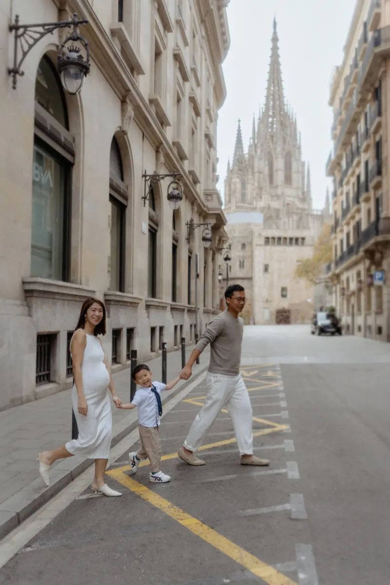 Smiling family holding hands and walking across a Barcelona street during a Vacation Portraits photoshoot near the cathedral.