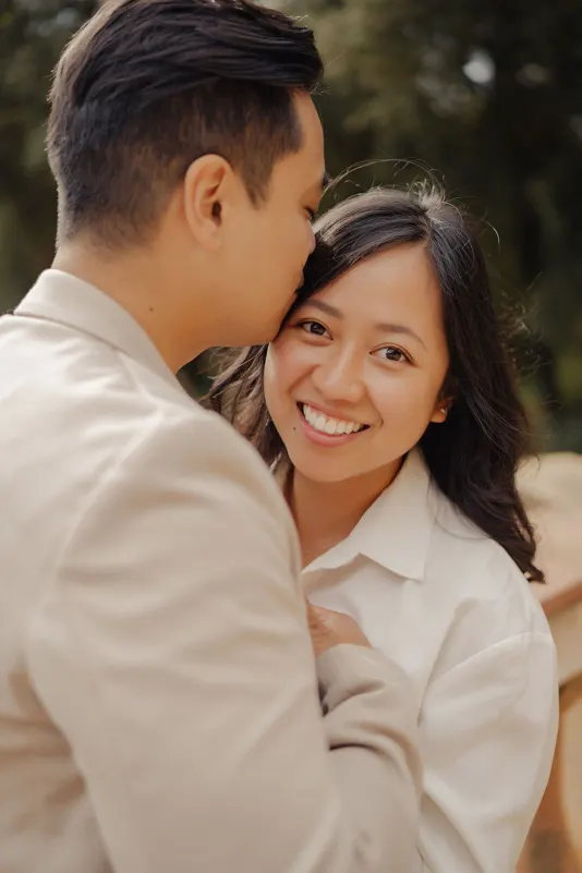 Smiling woman embraces her partner as he kisses her forehead during a couples & engagements photoshoot outdoors.