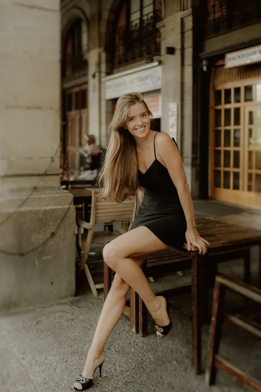 Smiling woman in a black dress poses at a café table during a Vacation Portraits photoshoot in a European city.