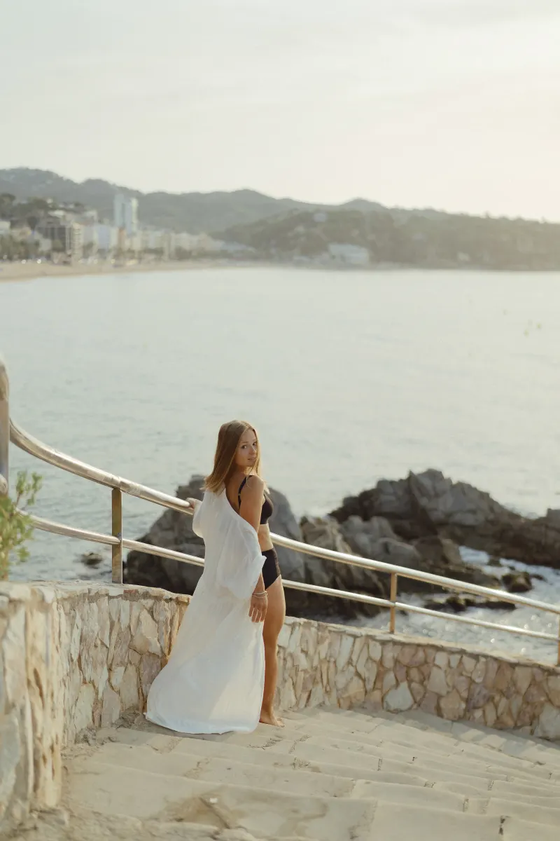 Smiling woman in a swimsuit and cover-up pauses on seaside steps during a Vacation Portraits session at sunset.