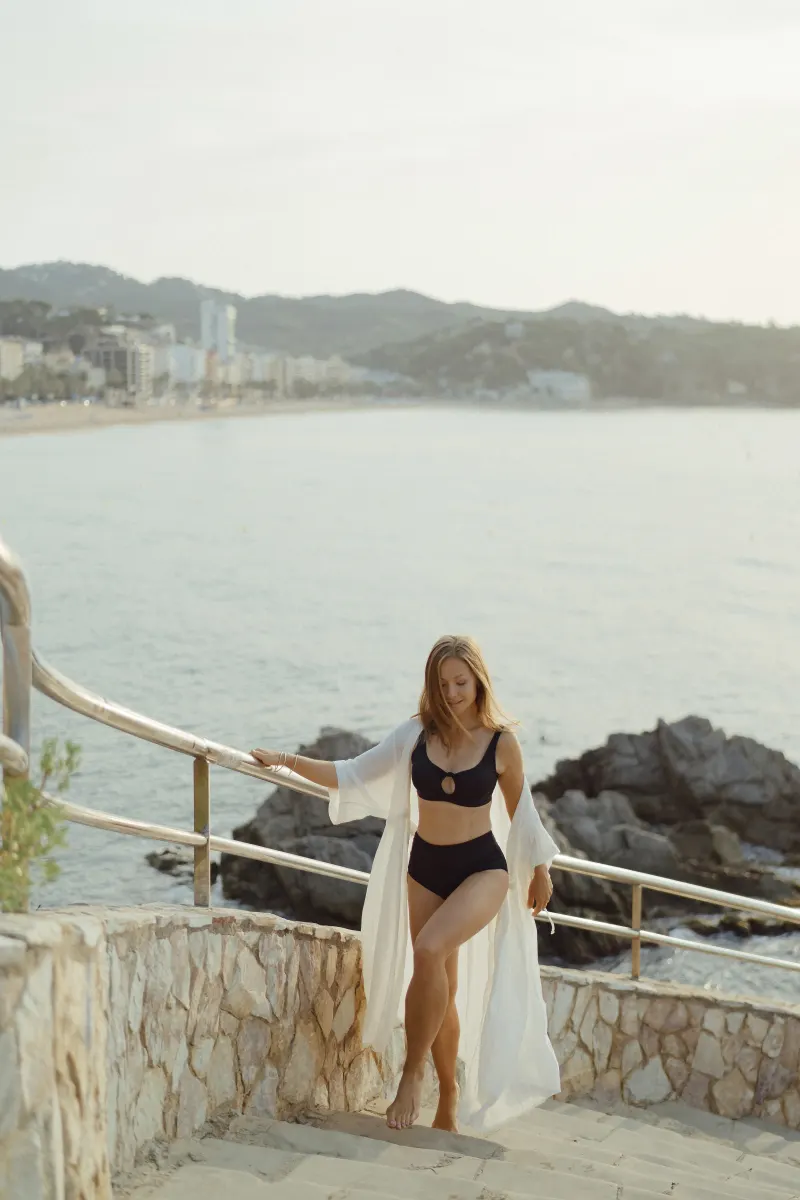 Smiling woman in swimwear walks up stone steps by the sea, captured during a Vacation Portraits session at sunset.