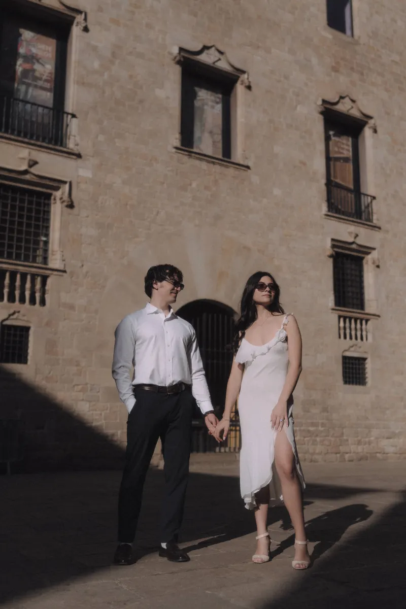 Stylish couple holding hands and enjoying the sunlight in front of a historic stone building during an engagement photoshoot.