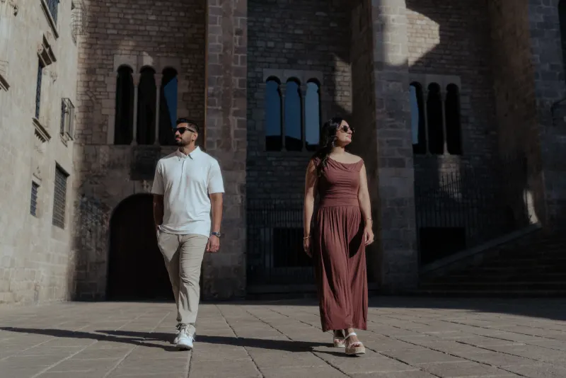 Stylish couple strolls confidently through a sunlit historic courtyard during their couples & engagements photoshoot.