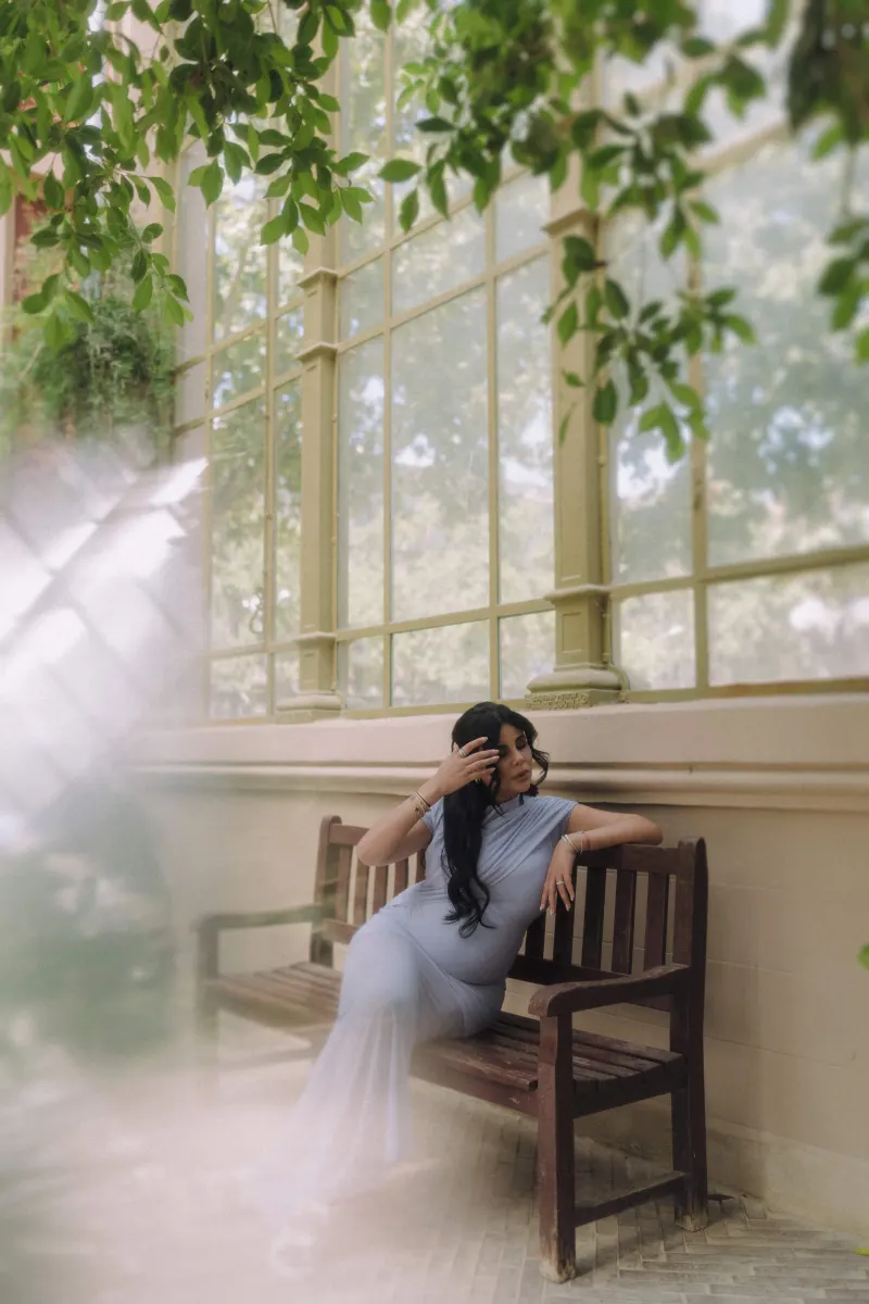 Stylish pregnant woman in a soft blue dress relaxes on a bench in a sunlit conservatory, Vacation Portraits session.