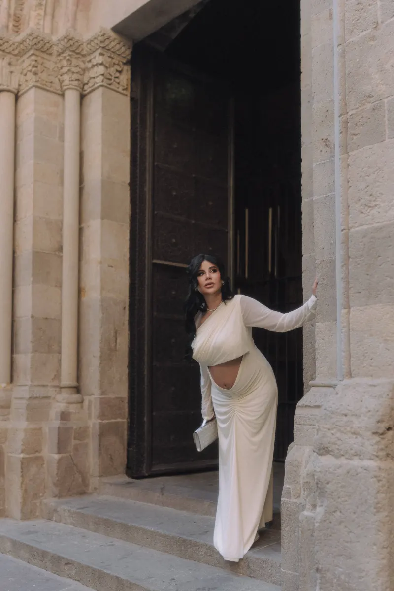 Stylish pregnant woman in a white gown poses gracefully in a historic stone doorway during a Vacation Portraits shoot.
