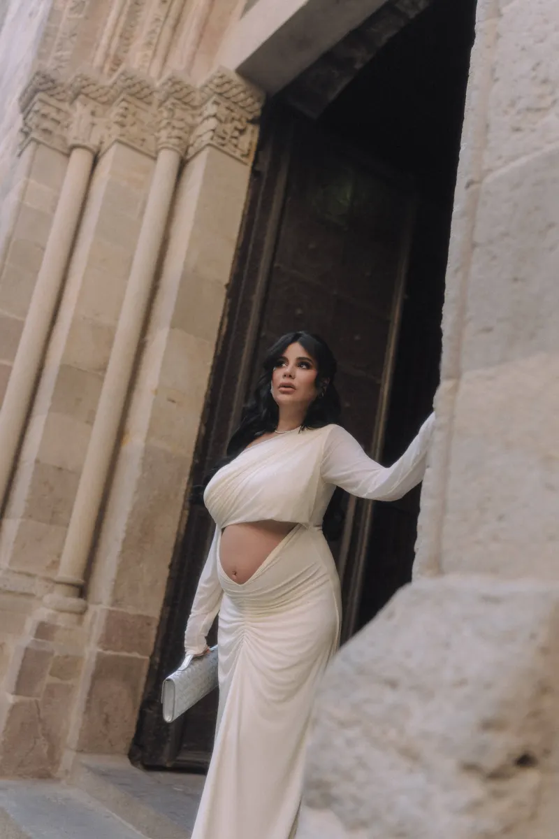 Stylish pregnant woman in a white gown stands by an ornate stone doorway during a Vacation Portraits photoshoot.