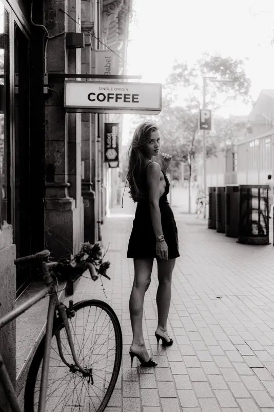 Stylish woman in a black dress glances back while walking past a café during a Vacation Portraits photoshoot.