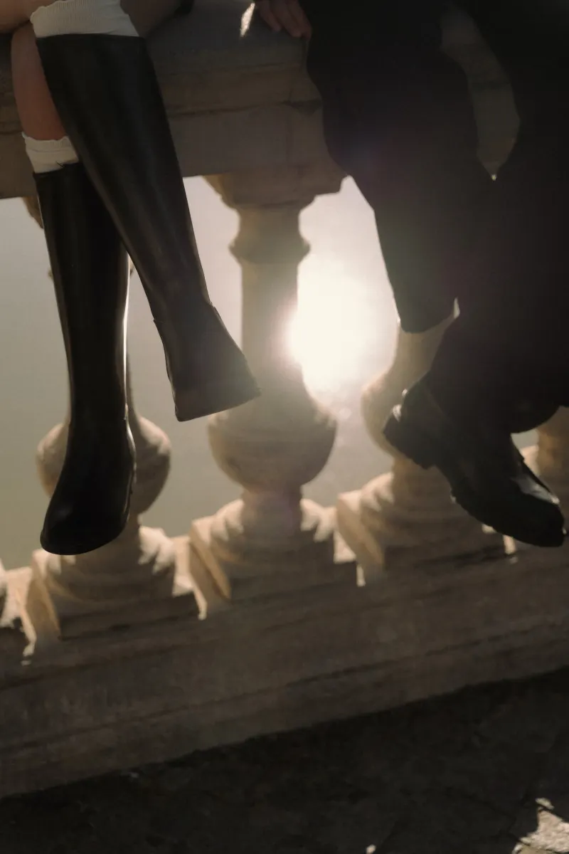 Sunlight glows behind a couple’s legs as they sit side by side on a stone railing during a Barcelona engagement shoot.