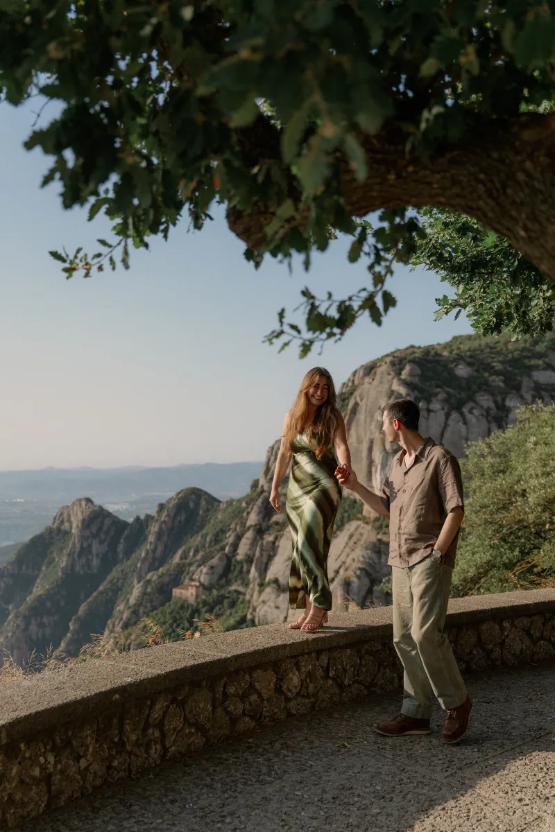 Tim and Megan hold hands beside the stone wall beneath leafy branches.
