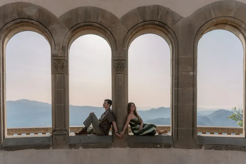 Tim and Megan sit in adjacent stone arches with mountains beyond.