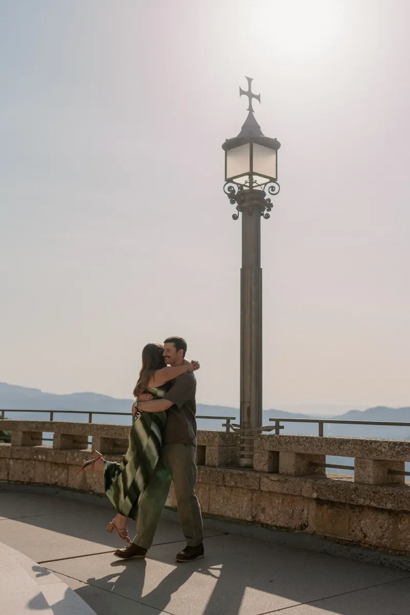 Tim lifts Megan into his arms on a sunny terrace beside a tall lamp.