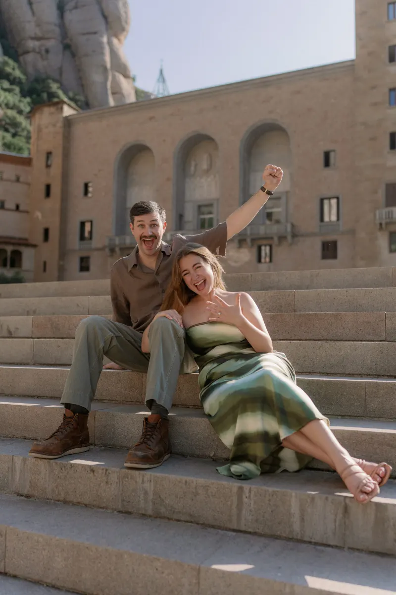 Tim raises his arm while Megan smiles and shows her engagement ring on the steps.
