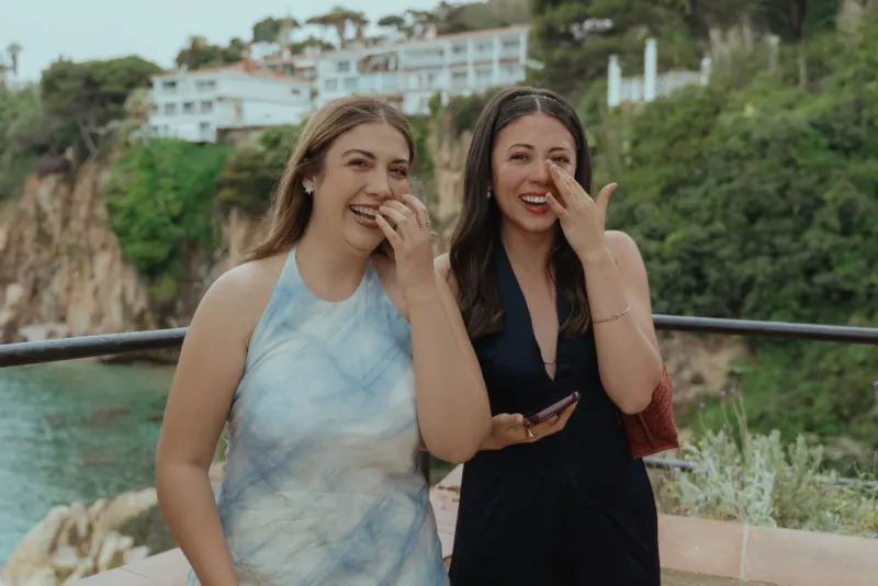 Two friends laugh and wipe away happy tears during a seaside surprise proposal photoshoot, surrounded by lush cliffs.