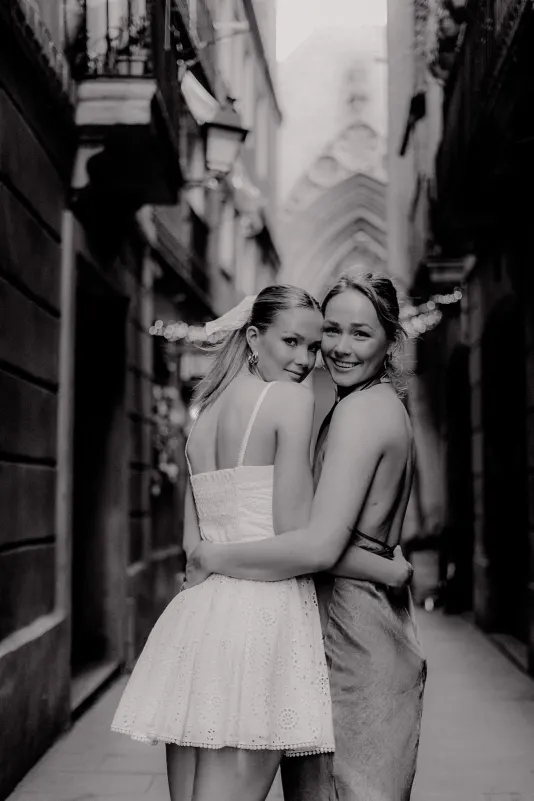 Two women embrace and smile in a narrow Barcelona street, Vacation Portraits photoshoot capturing a candid friendship moment.