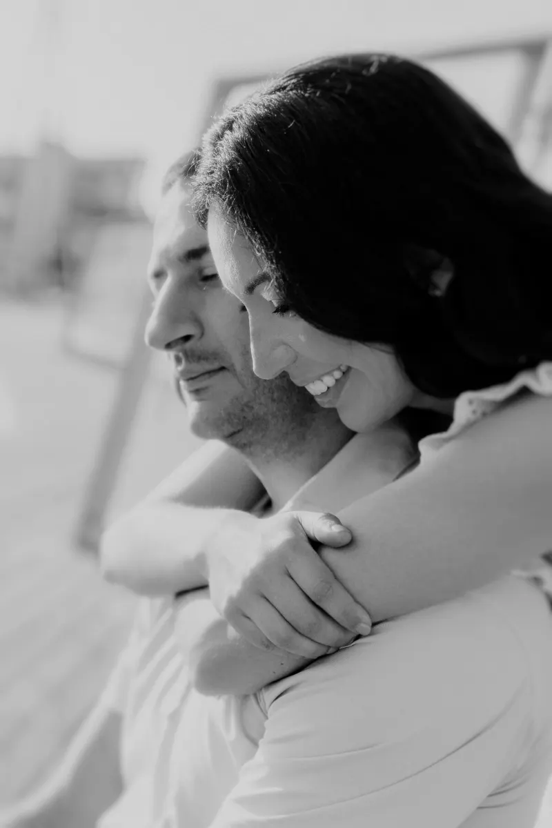 Woman embraces her partner from behind, both smiling softly during a couples & engagements photoshoot on a sunny dock.