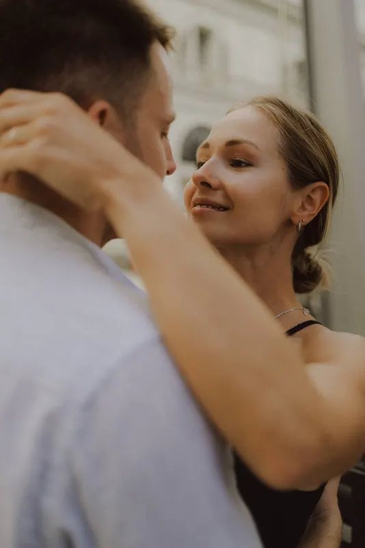 Woman gazes lovingly at her partner, arms around his neck, during a couples & engagements photoshoot outdoors.