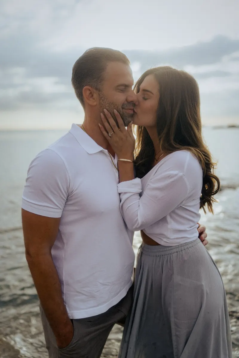 Woman gently kissing her partner’s cheek by the ocean, capturing a tender moment during a couples & engagements photoshoot.