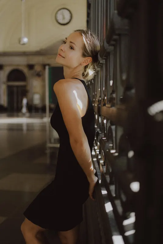 Woman in a black dress leaning against ornate railing, softly smiling during a couples & engagements session indoors.