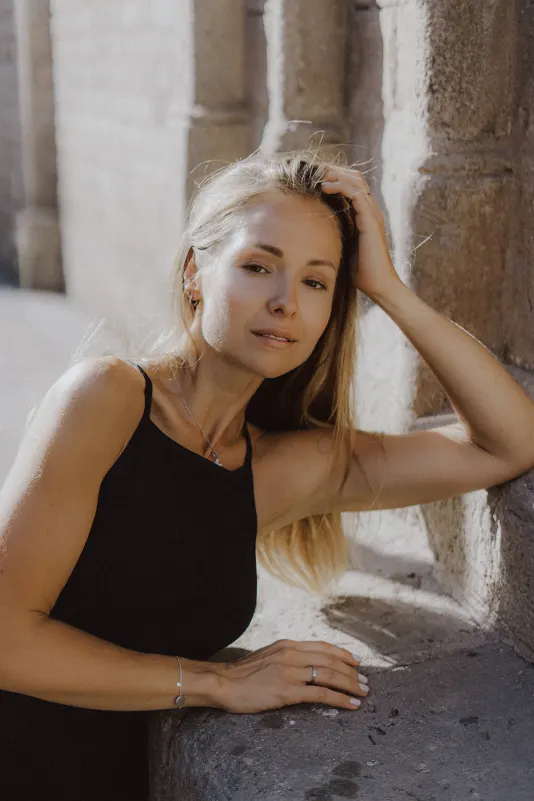 Woman in a black dress leans against a sunlit stone wall, sharing a quiet, thoughtful moment during an engagement shoot.