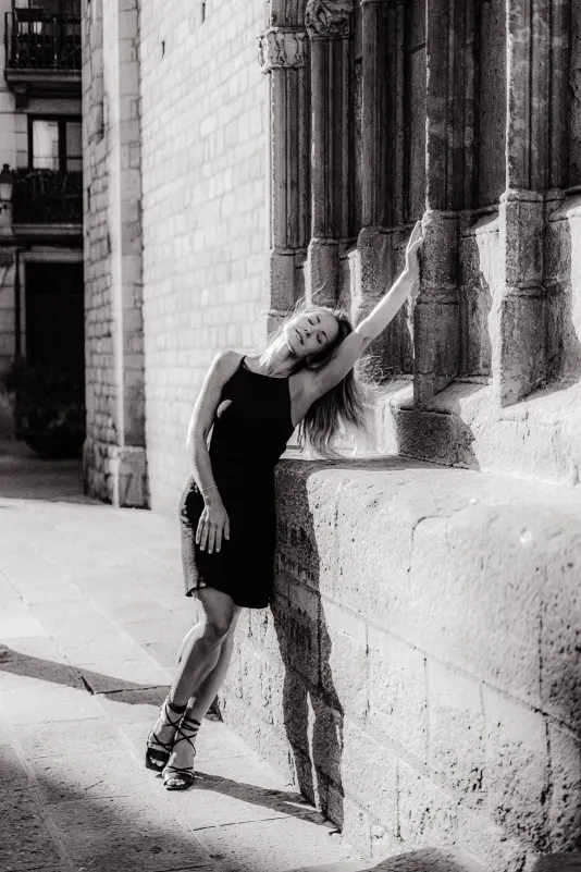 Woman in a black dress leans gracefully against a sunlit stone wall, enjoying a quiet moment during an engagement photoshoot.