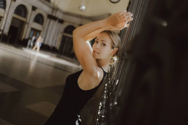 Woman in a black dress poses gracefully by an iron railing, sunlight streaming through a grand indoor space.