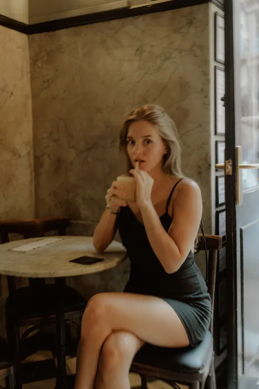 Woman in a black dress sips a drink at a marble café table during a relaxed Vacation Portraits photoshoot in Europe.