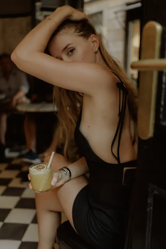 Woman in a black dress sits at a café doorway, holding a cocktail, during a relaxed Vacation Portraits photoshoot in Europe.