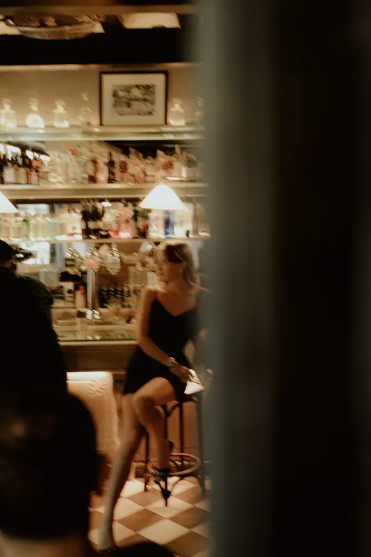 Woman in a black dress sits at a cozy bar, captured candidly during a Vacation Portraits photoshoot abroad.
