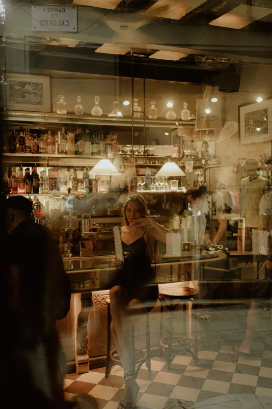 Woman in a black dress sits at a cozy bar, playfully holding a fan during a Vacation Portraits photoshoot in Europe.