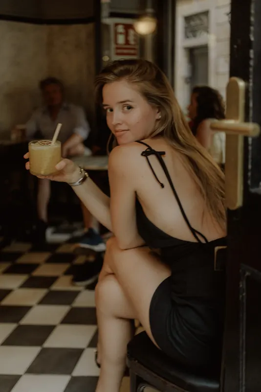 Woman in a black dress smiles back at the camera, holding a cocktail during a Vacation Portraits shoot in a cozy café.