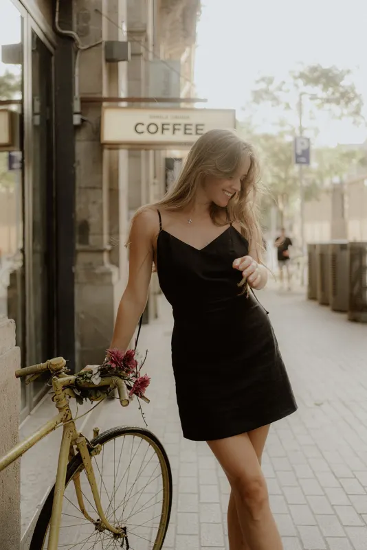 Woman in a black dress smiles beside a vintage bike with flowers during a Vacation Portraits shoot on a city street.