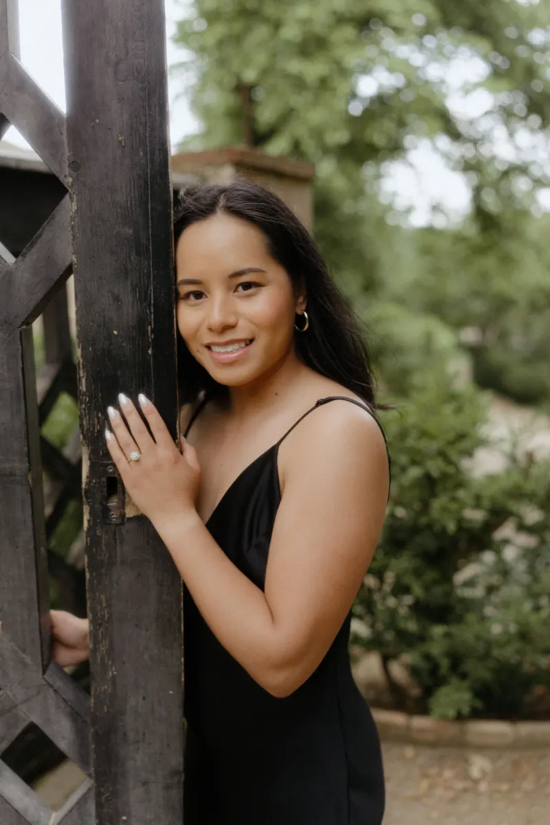 Woman in a black dress smiles by a garden gate, showing her engagement ring during a surprise proposal photoshoot.