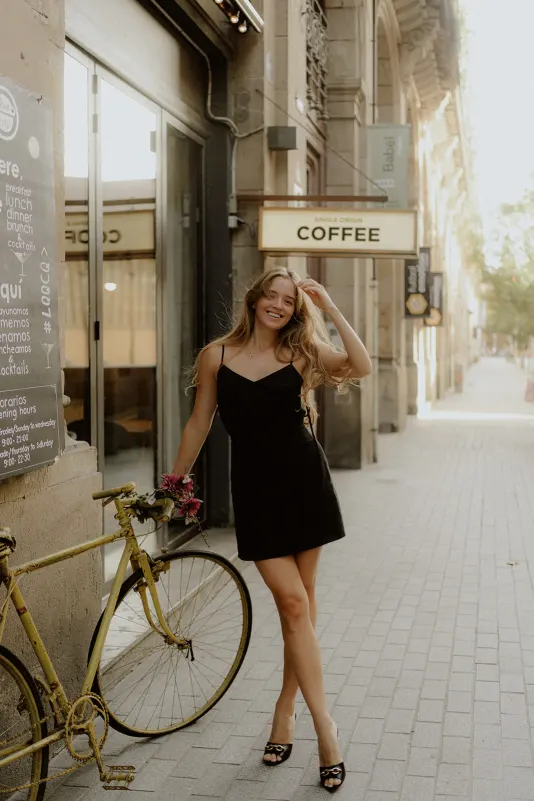 Woman in a black dress smiles by a vintage yellow bike outside a café during a Vacation Portraits photoshoot in Europe.