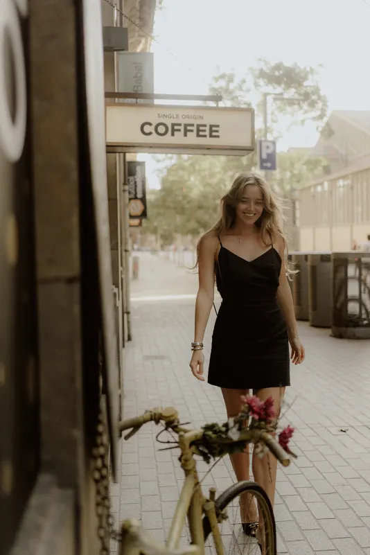 Woman in a black dress smiles while walking past a vintage bike outside a café during a Vacation Portraits shoot.