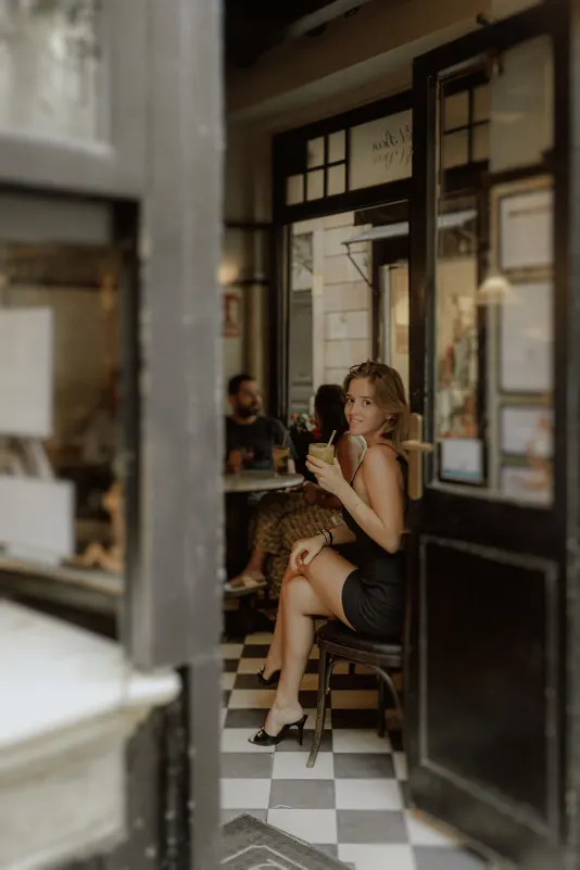 Woman in a black dress smiles with a cocktail at a cozy café, captured during a Vacation Portraits photoshoot in Europe.