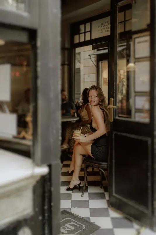 Woman in a black dress smiles with a drink at a cozy café, captured during a Vacation Portraits photoshoot in Europe.