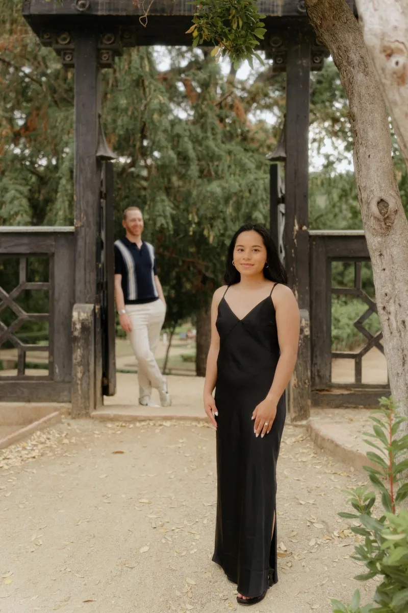 Woman in a black dress stands in a serene garden, her partner smiling in the background during a surprise proposal photoshoot