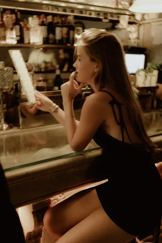 Woman in a black dress thoughtfully reads a menu at a cozy bar during a Vacation Portraits photoshoot in Europe.