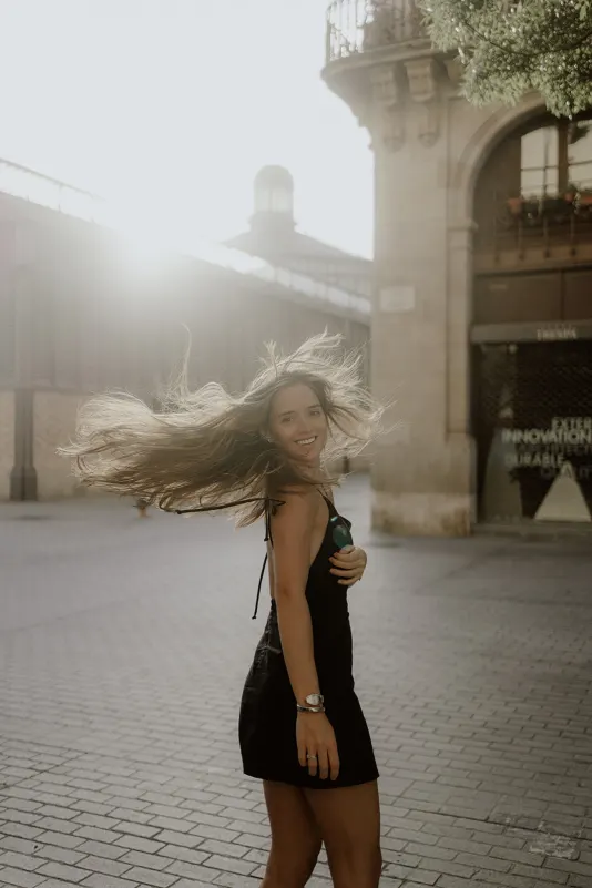 Woman in a black dress twirls joyfully on a sunlit European street during a Vacation Portraits photoshoot.