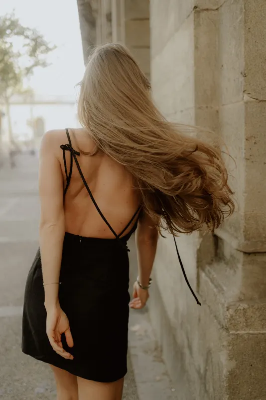 Woman in a black dress walks along a sunlit city street, hair flowing, during a Vacation Portraits photoshoot.