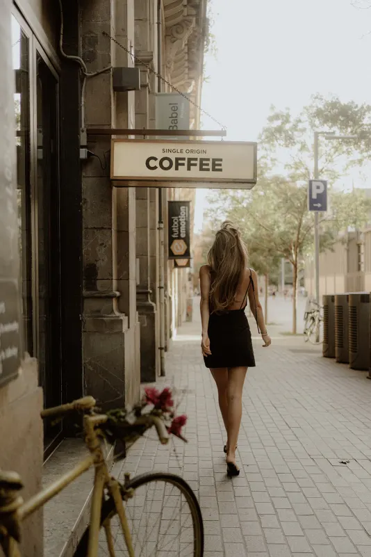 Woman in a black dress walks past a coffee shop and vintage bike, captured during a Vacation Portraits photoshoot.