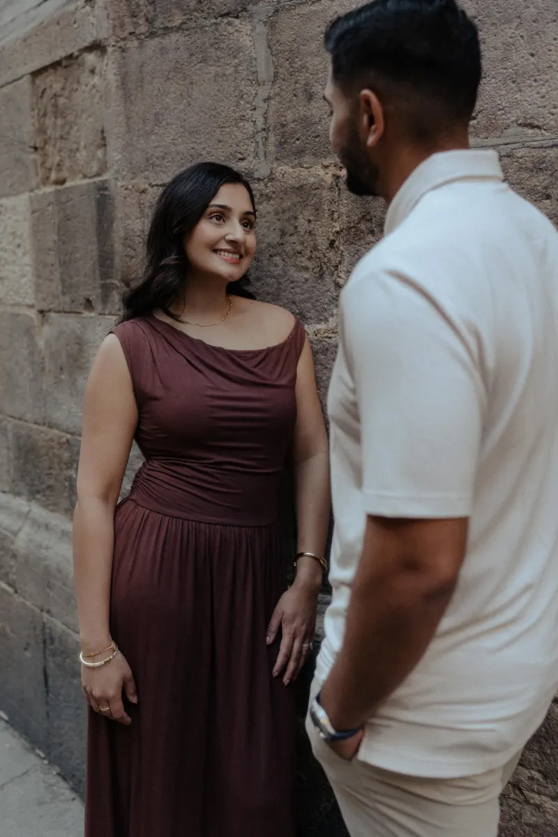 Woman in a burgundy dress smiles warmly at her partner during a couples & engagements photoshoot by a stone wall.