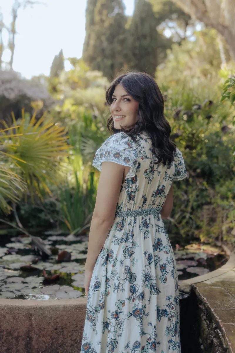Woman in a floral dress smiles over her shoulder by a tranquil pond during a surprise proposal photoshoot in a garden.