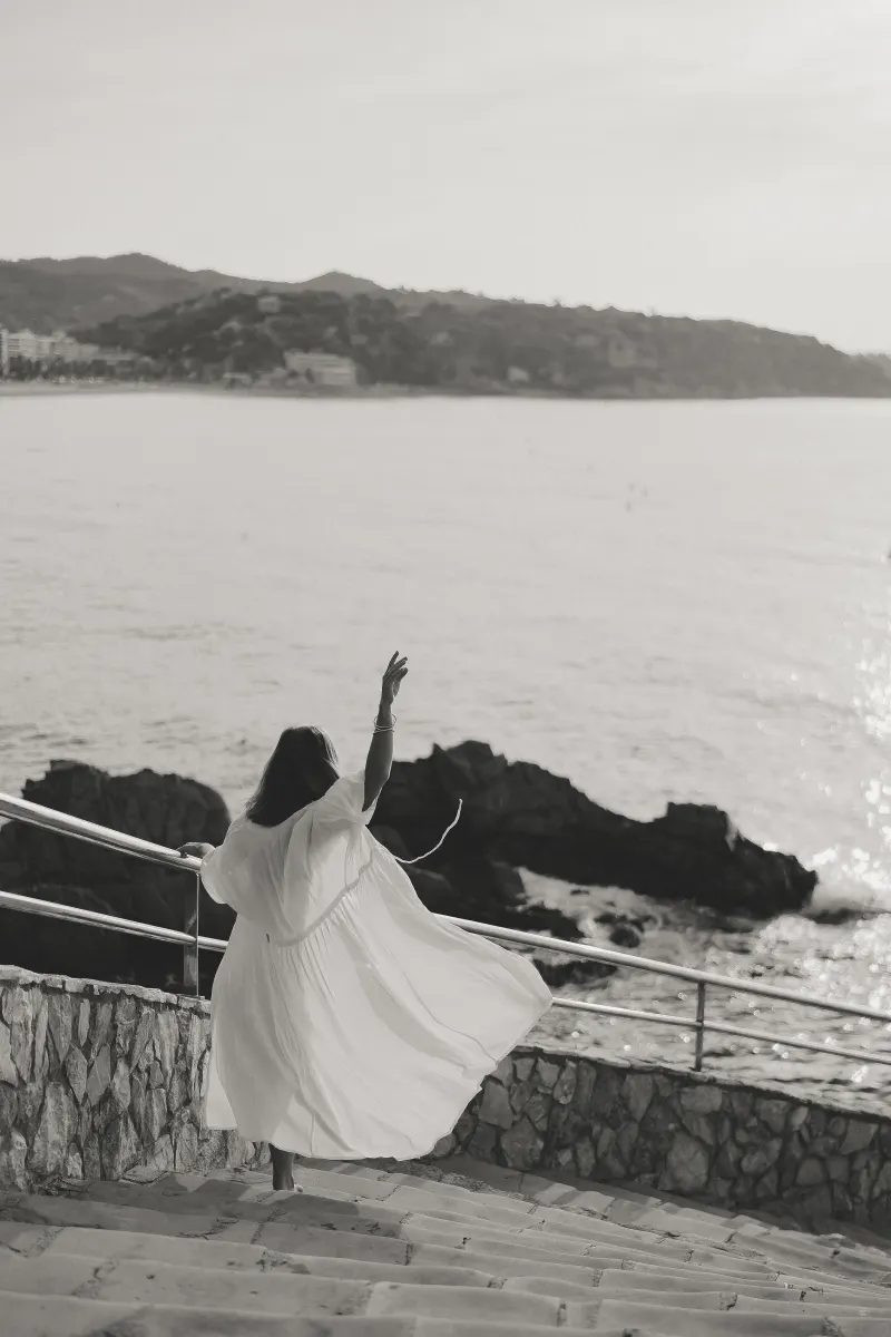 Woman in a flowing dress twirls on seaside steps, celebrating a carefree moment during a Vacation Portraits photoshoot.