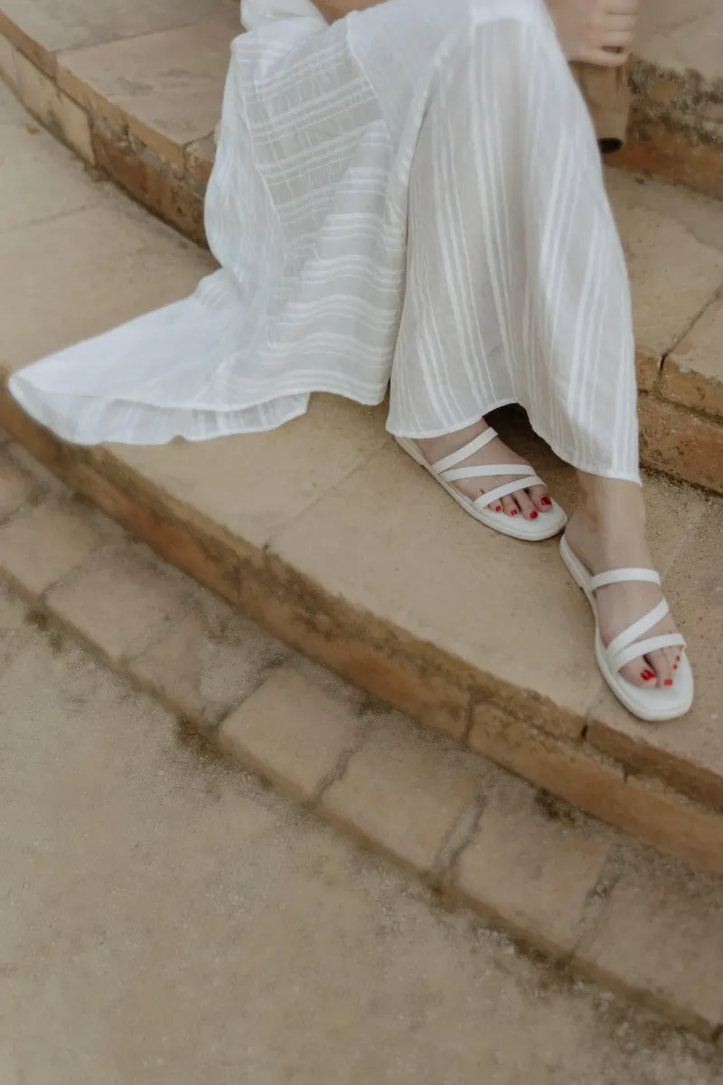 Woman in a flowing white dress and sandals sits on stone steps, captured during a couples & engagements photoshoot.