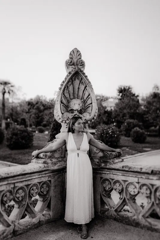 Woman in a flowing white dress leans on ornate stone railing, captured during a romantic couples photoshoot in the garden.
