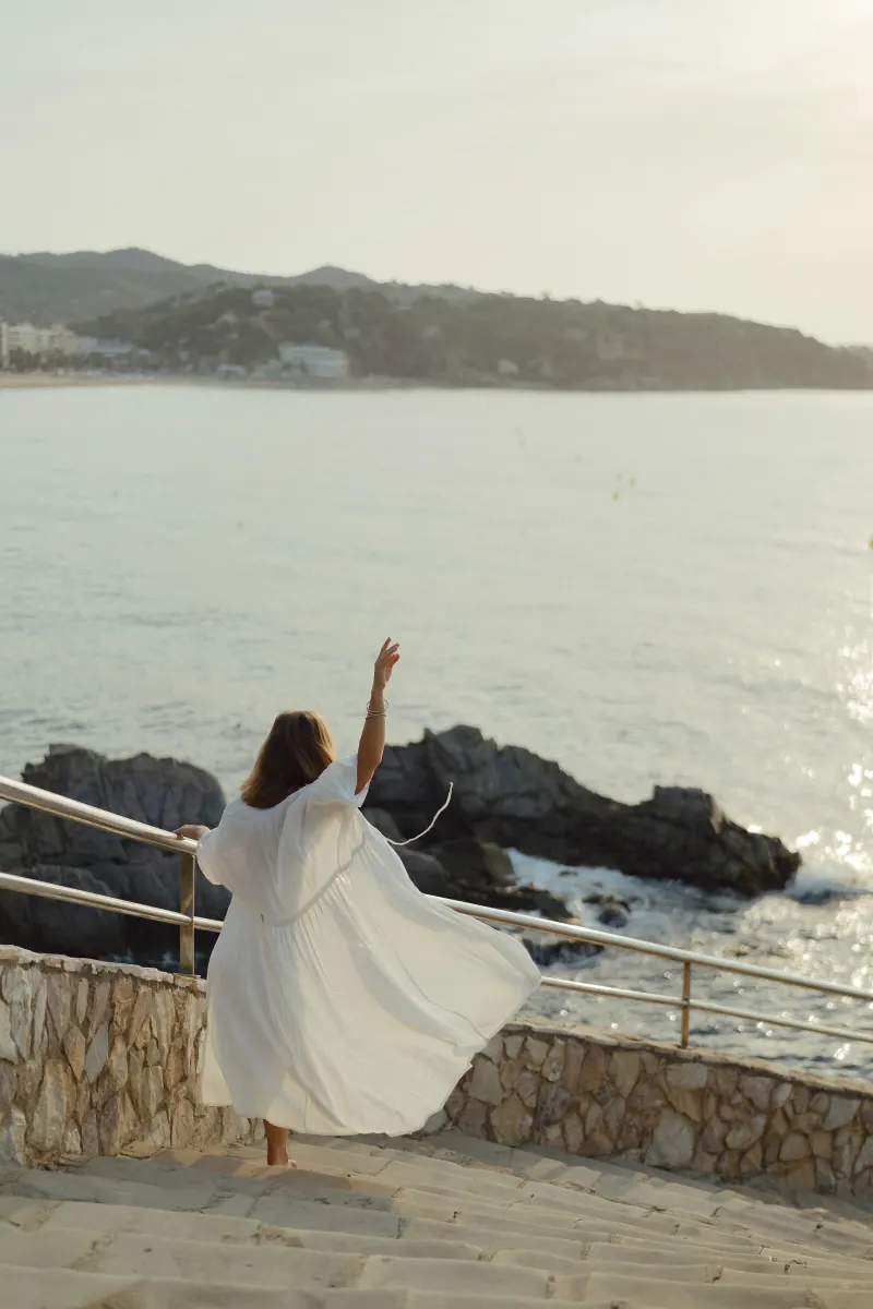 Woman in a flowing white dress twirls on seaside steps, celebrating golden hour during a Vacation Portraits photoshoot.