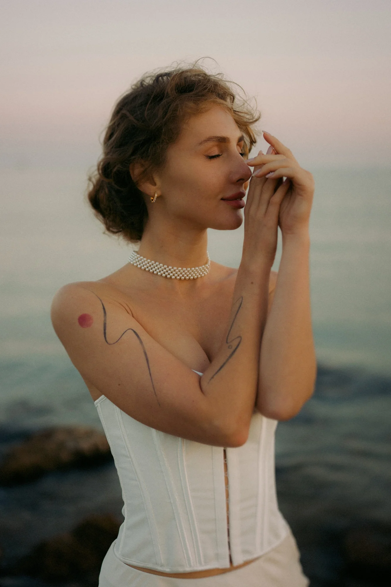 Woman in a white corset stands by the sea at sunset, eyes closed in a peaceful moment during a Vacation Portraits session.