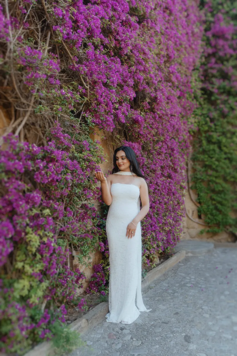 Woman in a white dress admires purple bougainvillea, captured during a romantic surprise proposal photoshoot.